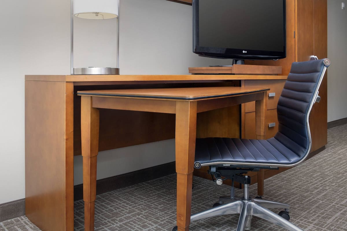 A hotel room with a wooden desk, a black office chair, a flat-screen TV, and a lamp.