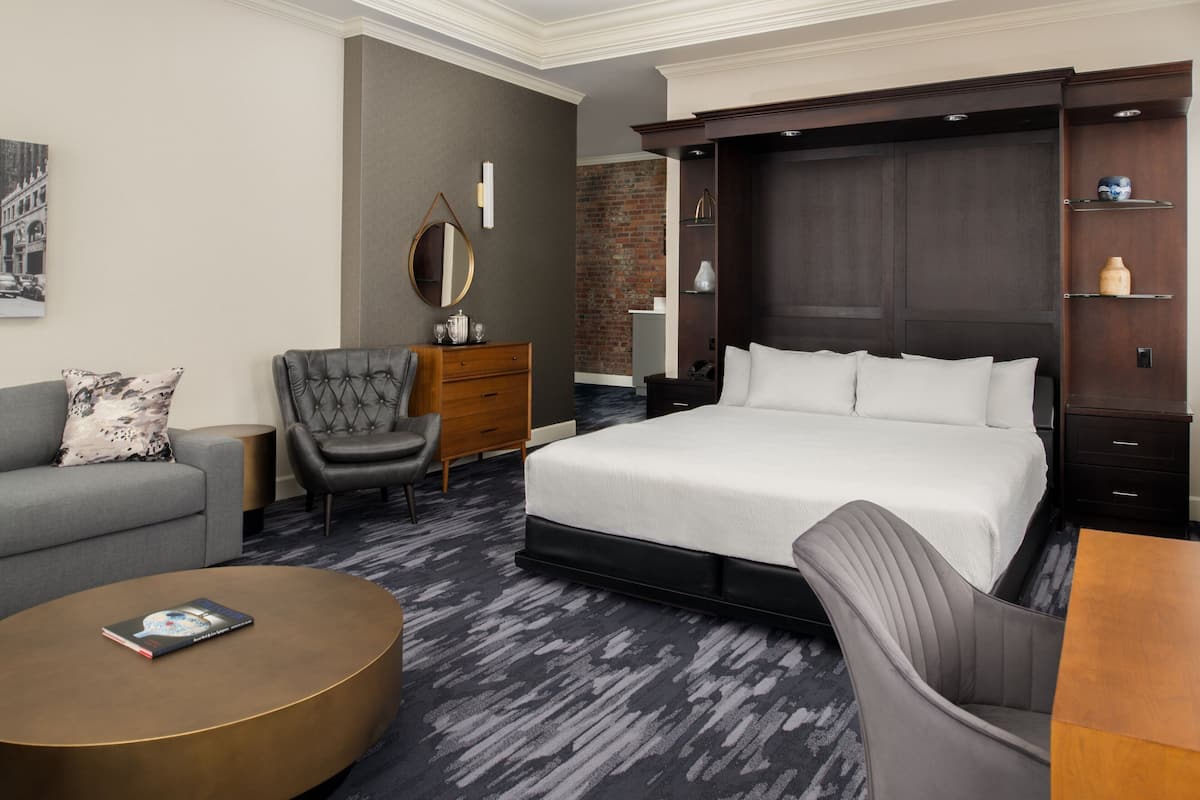 Hotelkamer met een groot bed, een grijze fauteuil, een houten nachtkastje, een ronde salontafel, een dressoir, een spiegel en een bakstenen muur.
