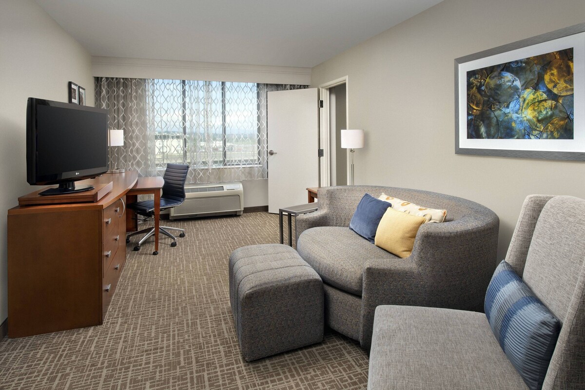 A hotel room with a flat-screen TV, a desk, a comfortable sofa, and a large window with a view.