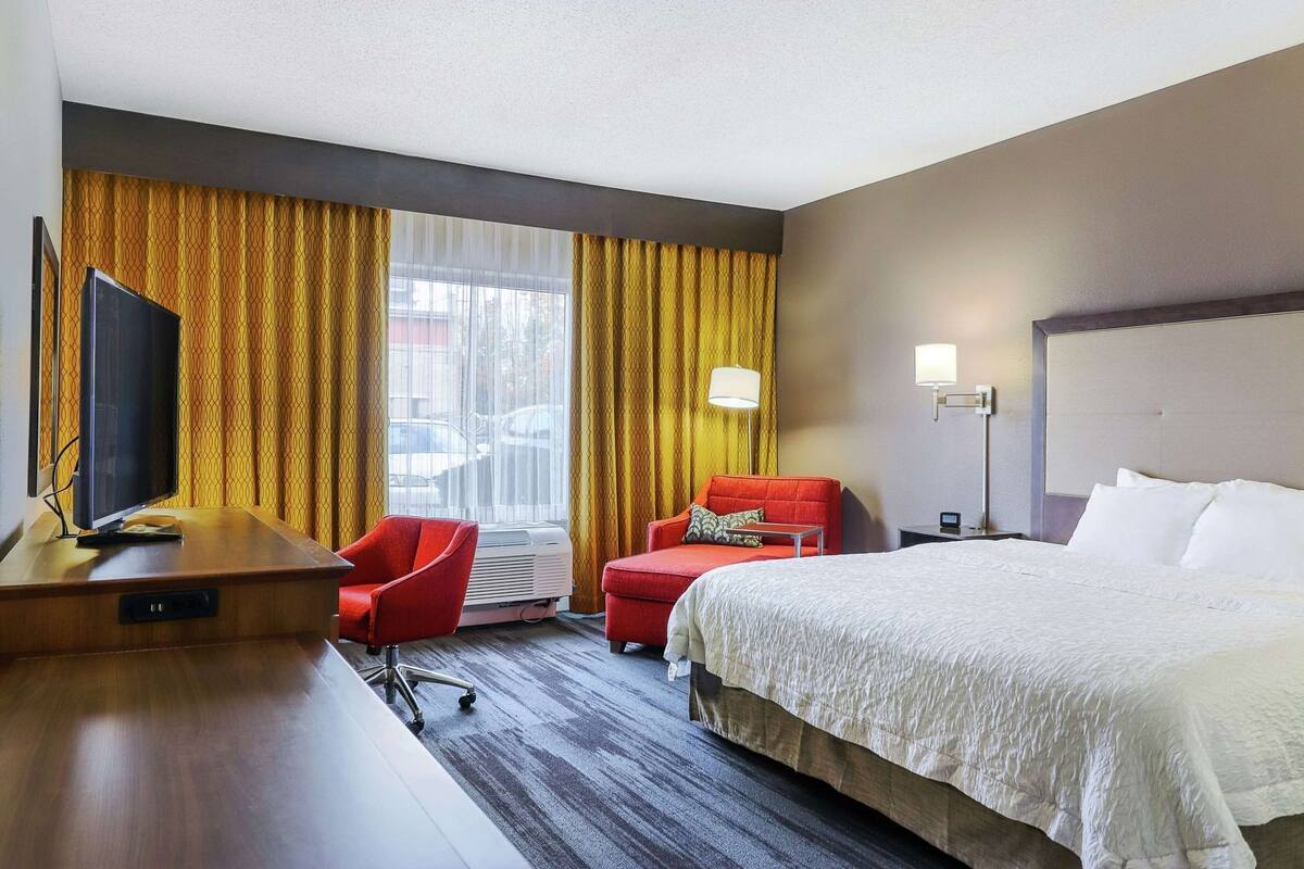 A hotel room with a bed, a desk with a TV, a red chair, a lamp, and a window with curtains.
