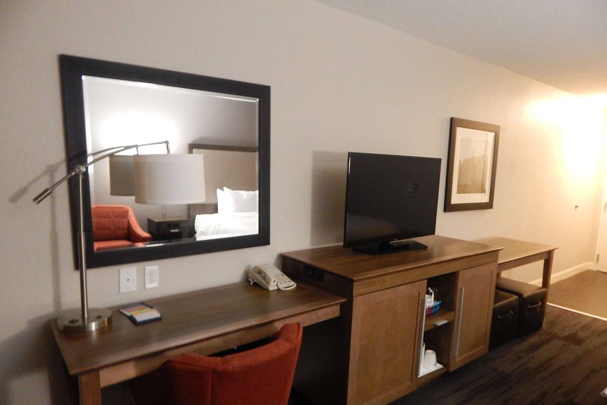A hotel room with a wooden desk, a television, a mirror, a telephone, and a red chair.