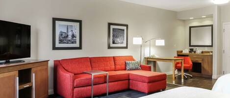 A hotel room with a bed, a red sofa, a desk with a chair, a TV, and framed pictures on the wall.