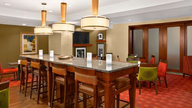 A hotel dining area with a central island, wooden chairs, and hanging pendant lights. - Hampton Inn Heath-Newark (Heath)