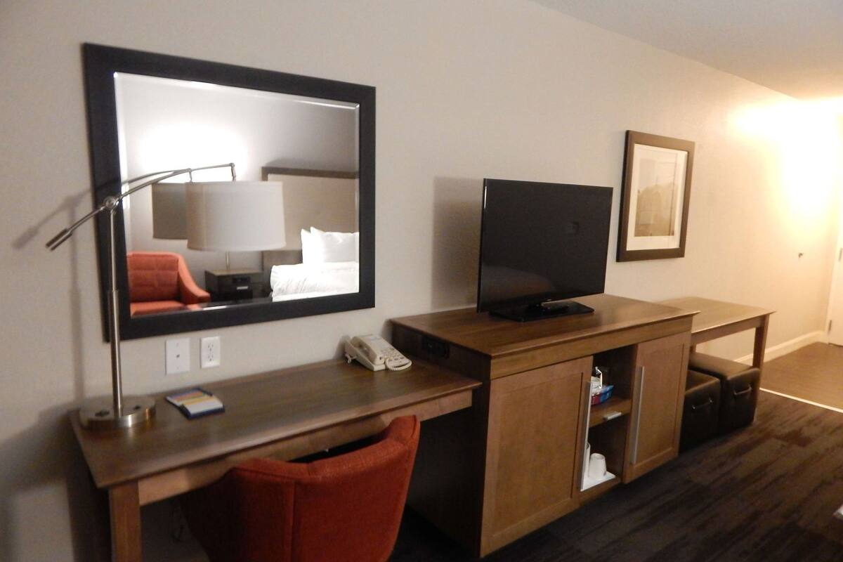 A hotel room with a wooden desk, a television, a mirror, a red chair, a telephone, and a framed picture on the wall.
