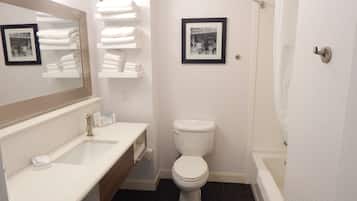 Een badkamer met een witte wastafel, toilet en bad. Handdoeken liggen op een plank gestapeld en er hangt een lijst met een afbeelding aan de muur.