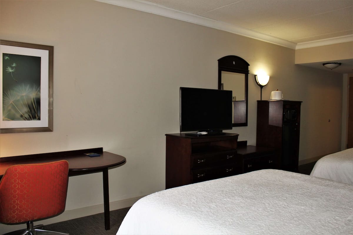 Quarto de hotel com cama, escrivaninha com cadeira, televisão, cômoda e espelho.