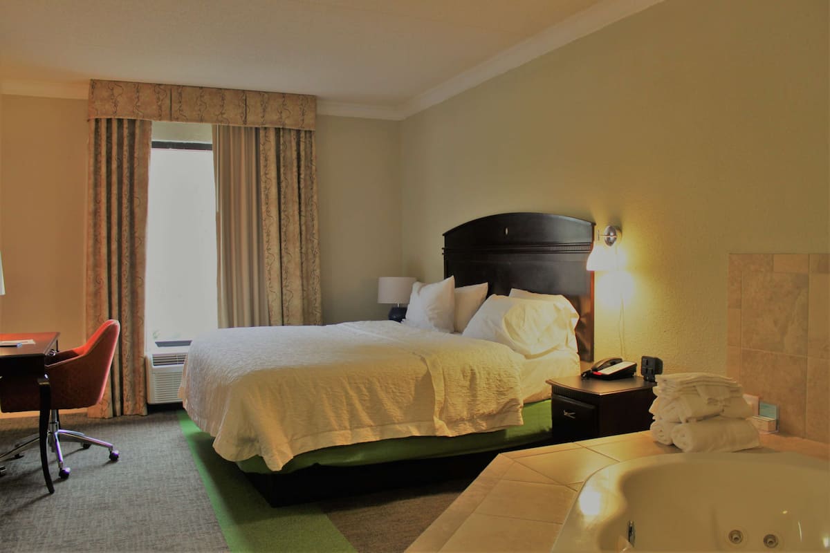 Quarto de hotel com cama, escrivaninha, cadeira, abajur, telefone, pilha de toalhas e banheira.