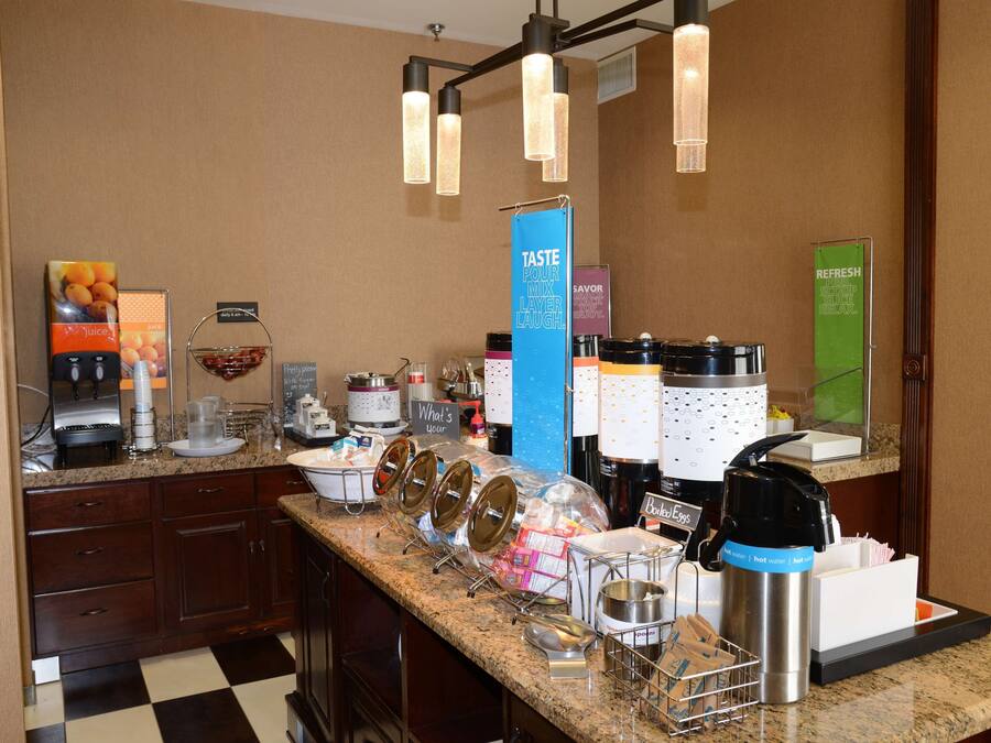 A hotel breakfast bar with various food and drink options, including juice dispensers, coffee makers, and a sign for a taste mixer layer.