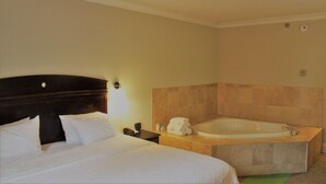 A hotel room with a large bed, a separate bathroom with a bathtub, and a small table with a phone and a bottle.