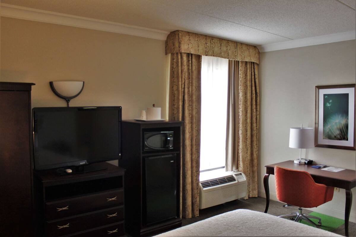Quarto de hotel com televisão de tela plana, mini-geladeira, escrivaninha com luminária, cadeira e uma grande janela com cortinas.