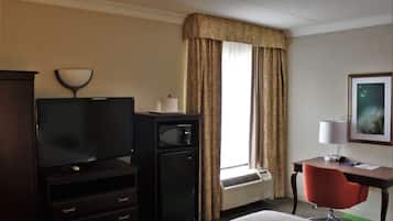 A hotel room with a flat-screen TV, a small refrigerator, a desk with a lamp, a chair, and a large window with curtains.