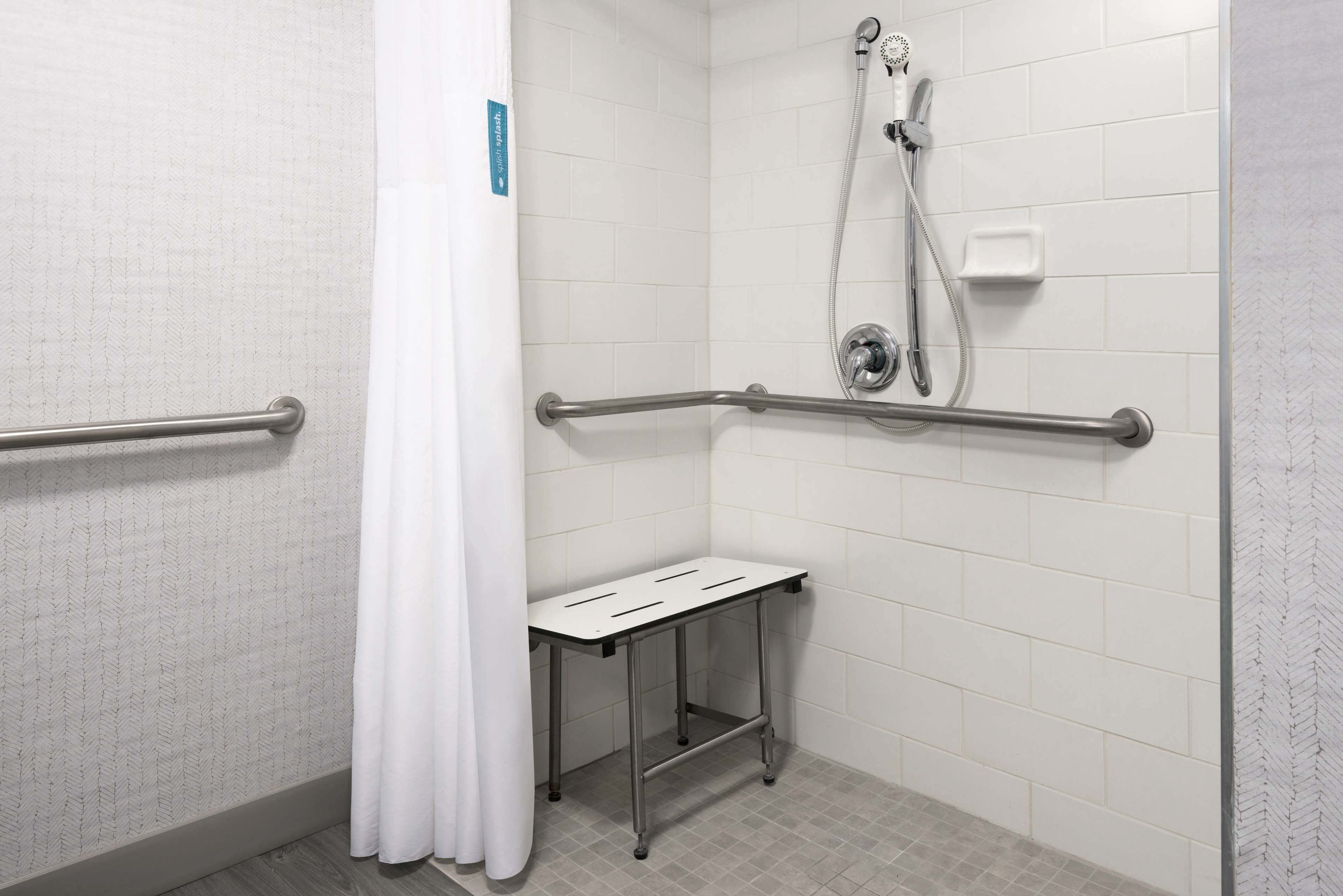 Área de duche com paredes azulejadas em branco, chuveiro, mangueira de duche, barra de apoio e um pequeno banco branco.