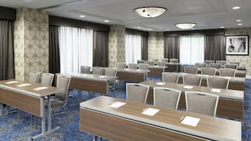 Sala de conferência com várias mesas e cadeiras, janelas amplas com cortinas e parede com padrão.
