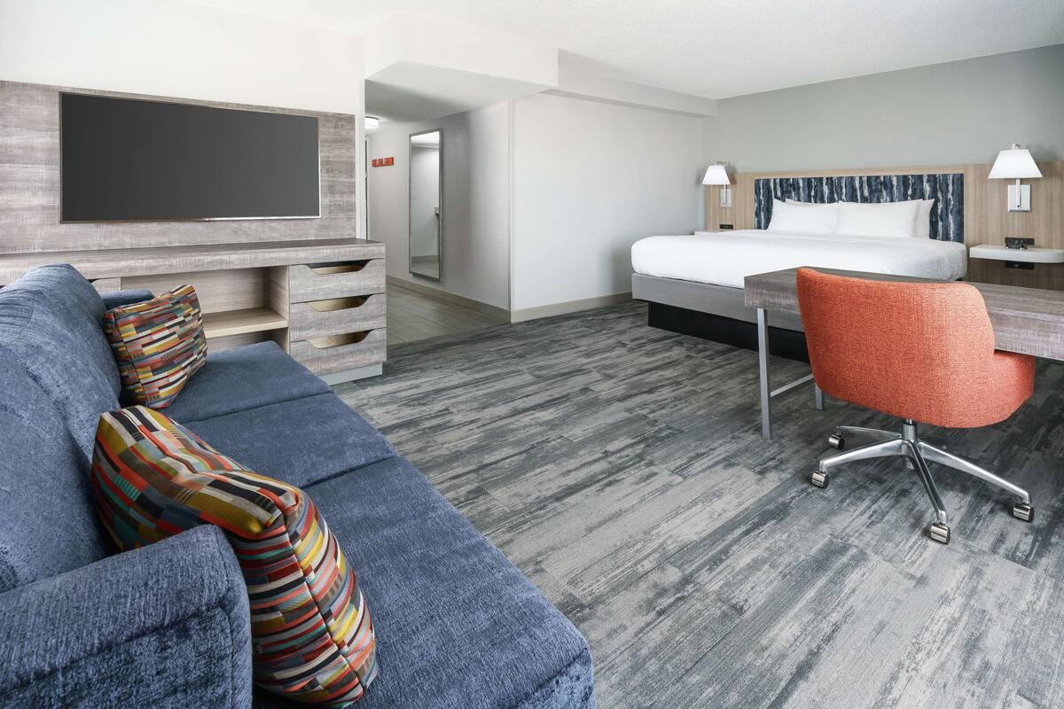 Een hotelkamer met een grote flatscreen televisie aan de muur, een blauwe bank met kleurrijke kussens, een bed met houten hoofdbord, een oranje bureaustoel, en een klein bureau met een lamp.