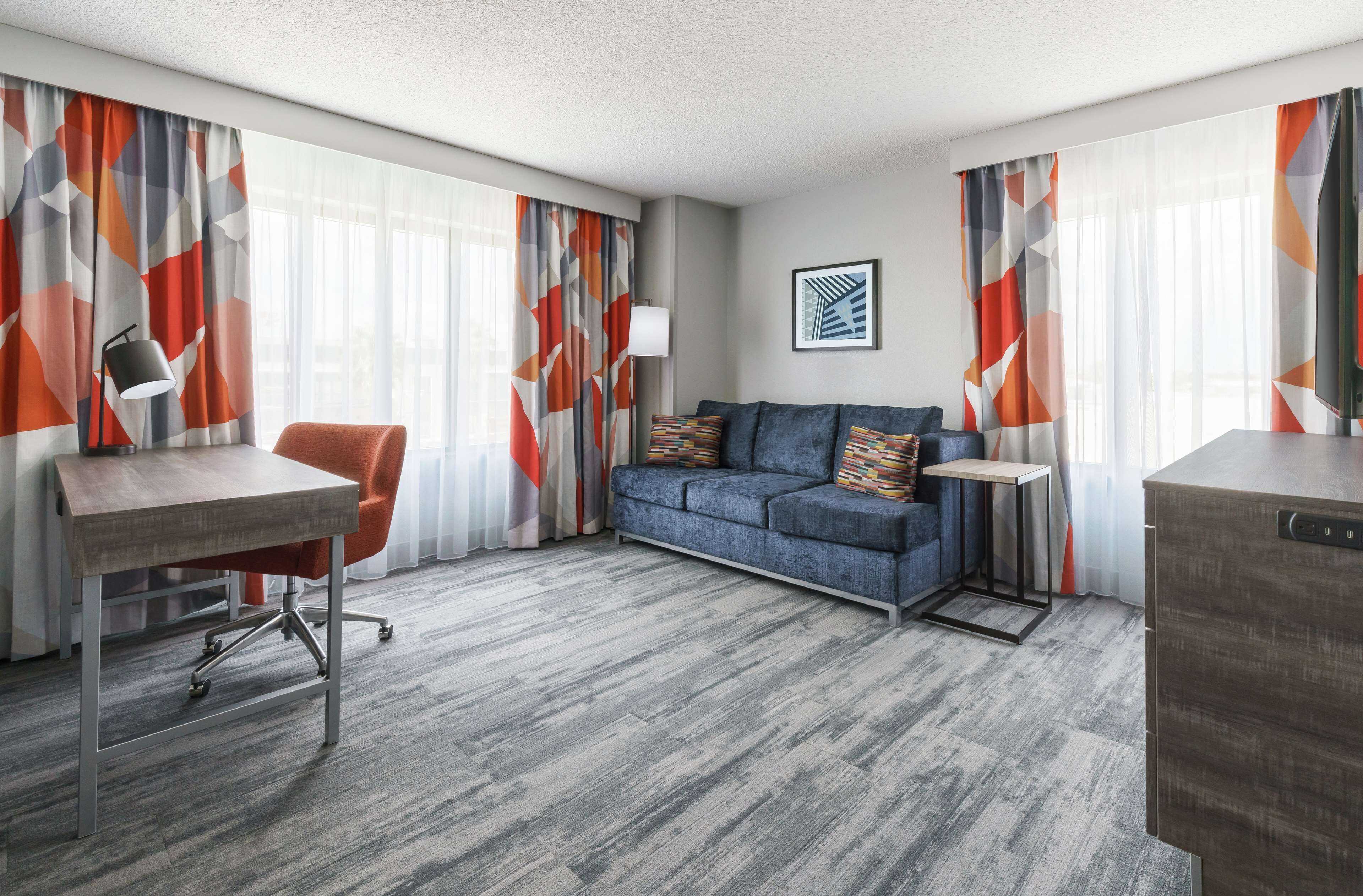Une chambre d’hôtel moderne avec un tapis gris, un canapé gris agrémenté de coussins colorés, un bureau en bois et une petite table d’appoint.