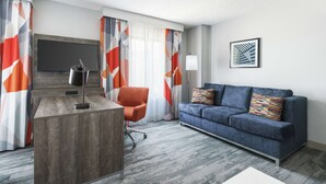 Een moderne hotelkamer met een houten bureau, een oranje stoel, een blauwe bank en een groot raam met geometrische gordijnen.