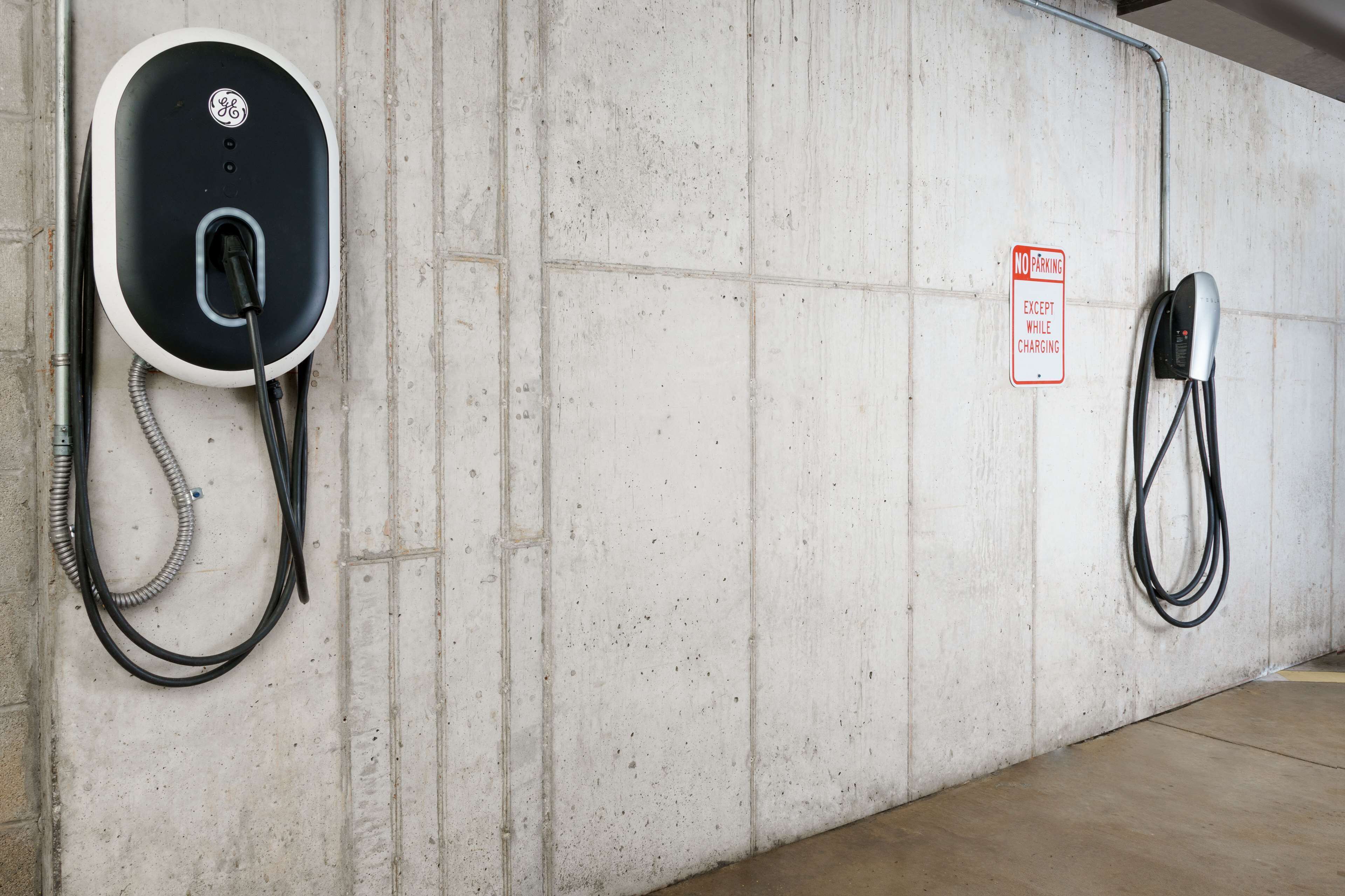 Un área de estacionamiento con estaciones de carga para vehículos eléctricos, un muro de concreto y un letrero que indica 'Prohibido estacionar excepto durante la carga'.
