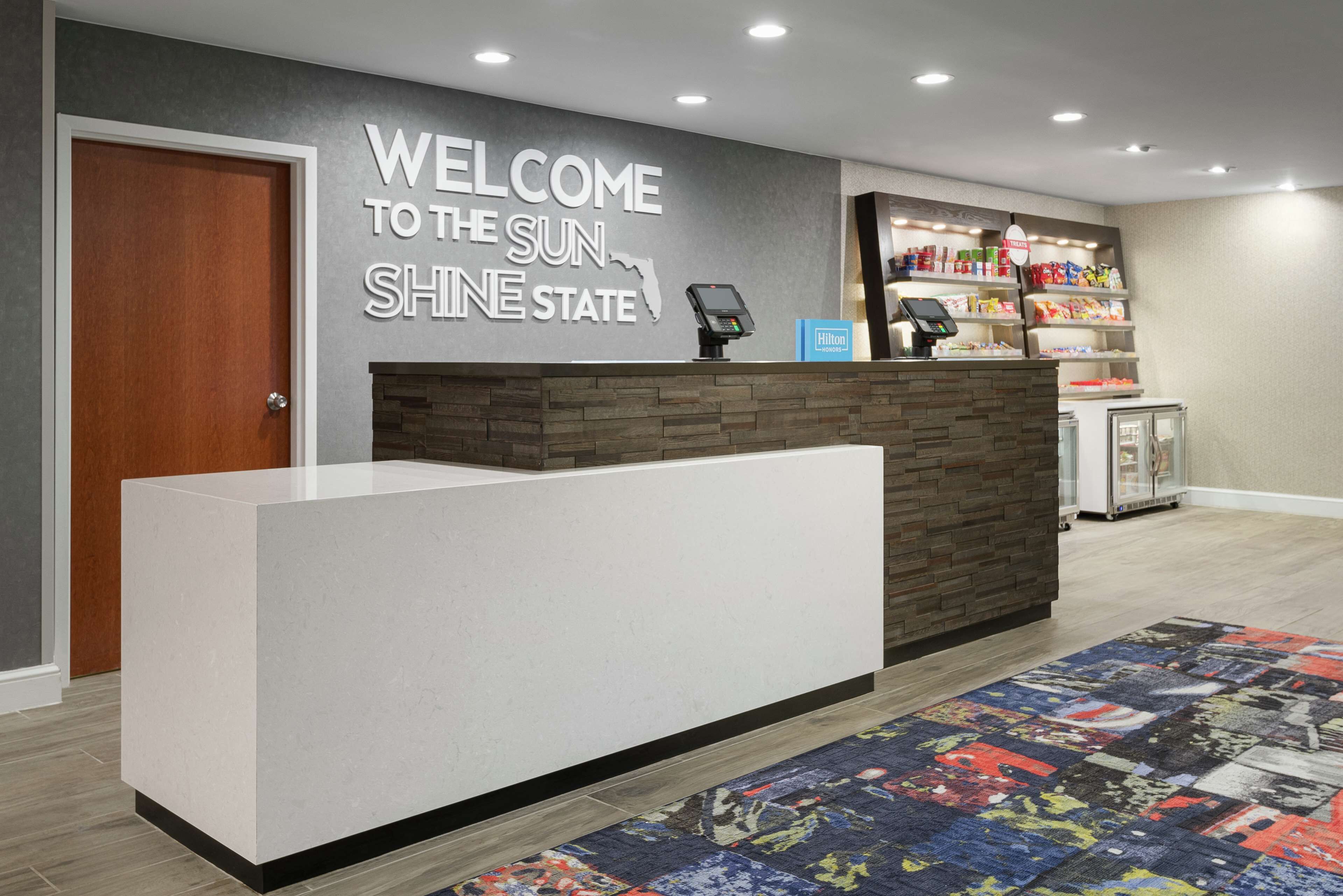 A hotel reception area with a modern design, a welcome sign, a self-service kiosk, and a minibar.