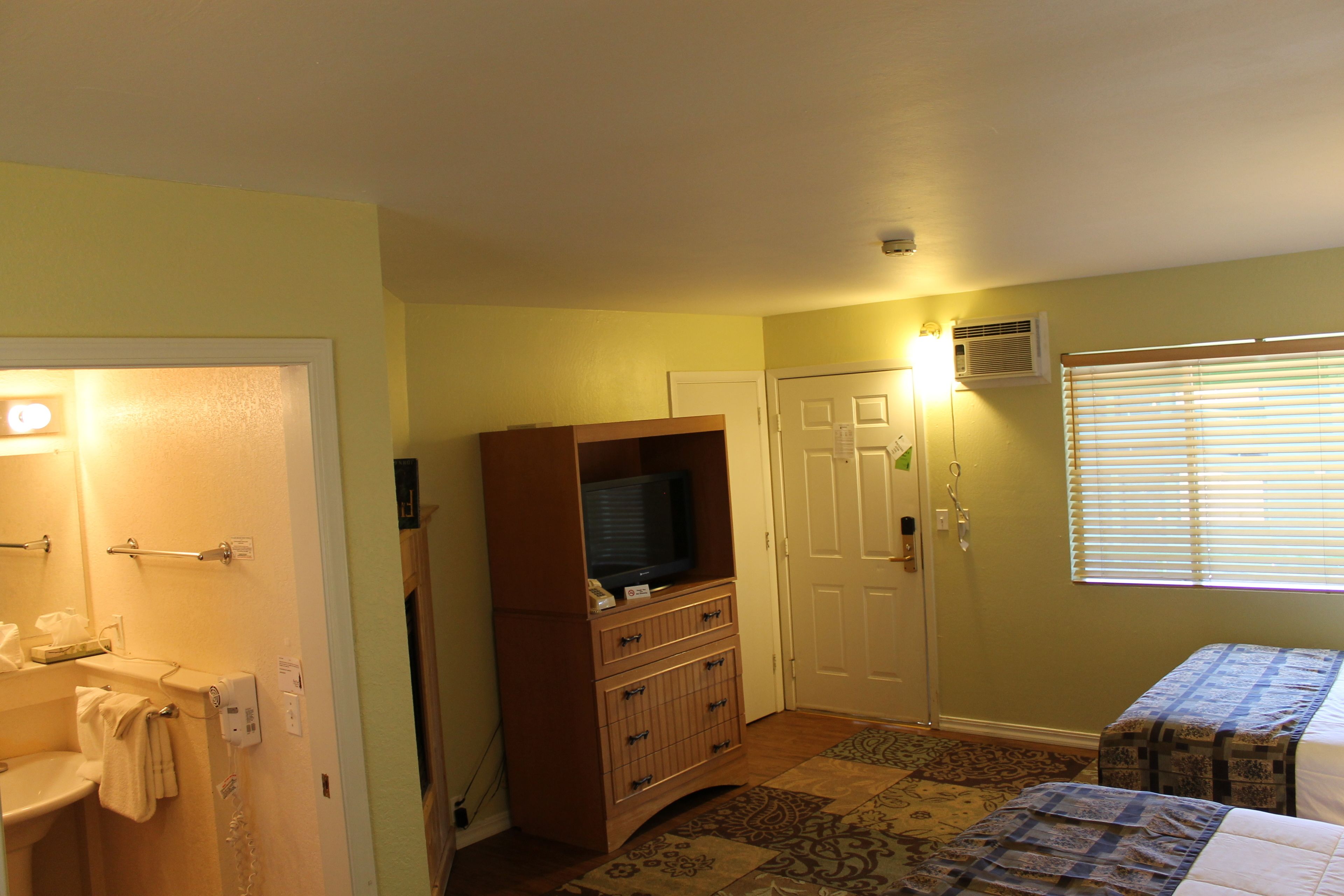 A bedroom with a bed, a television on a wooden cabinet, a bathroom with a toilet and sink, and a window with blinds.