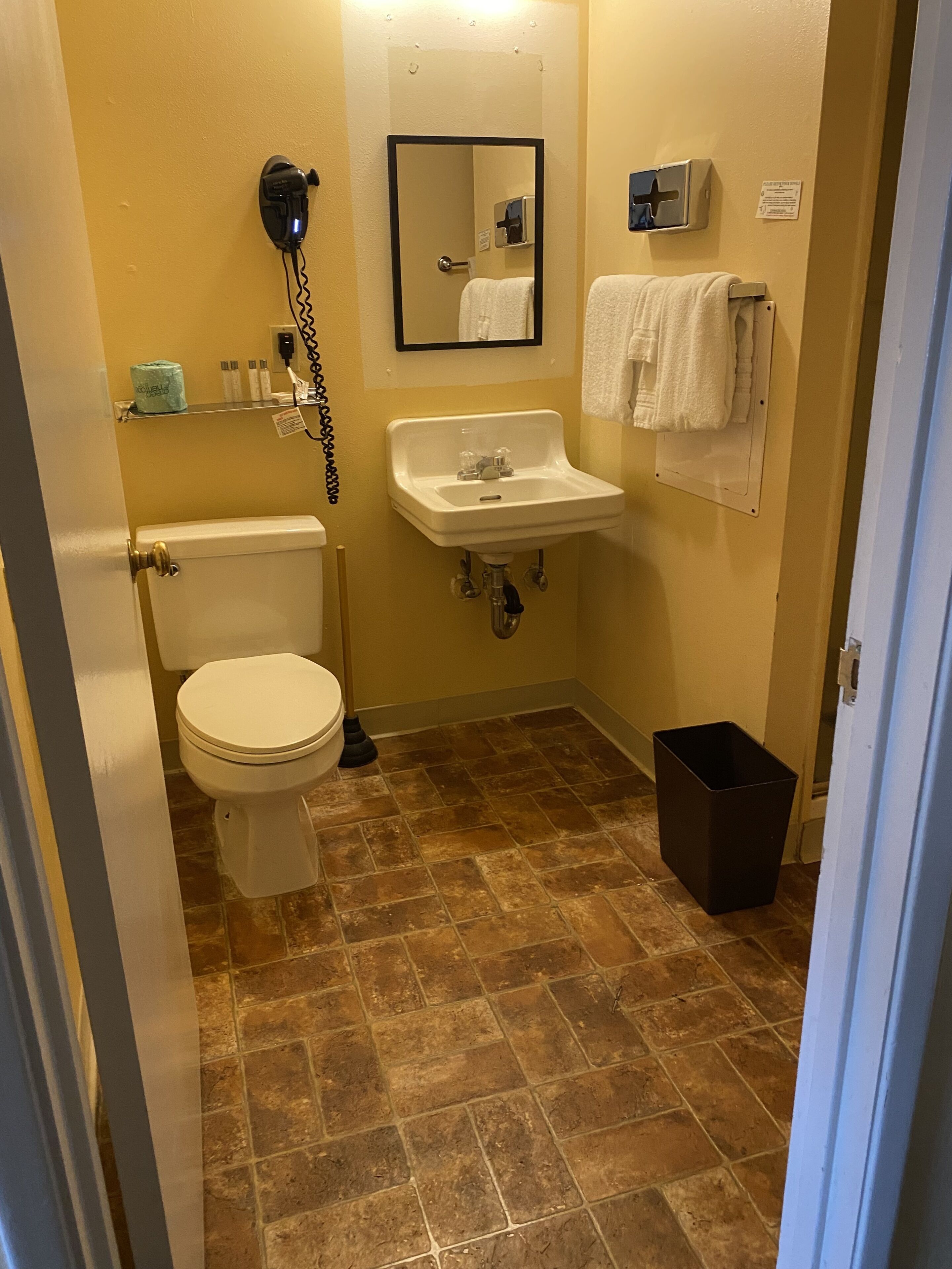 A bathroom with a toilet, sink, and mirror. There is a towel rack with towels and a trash can.