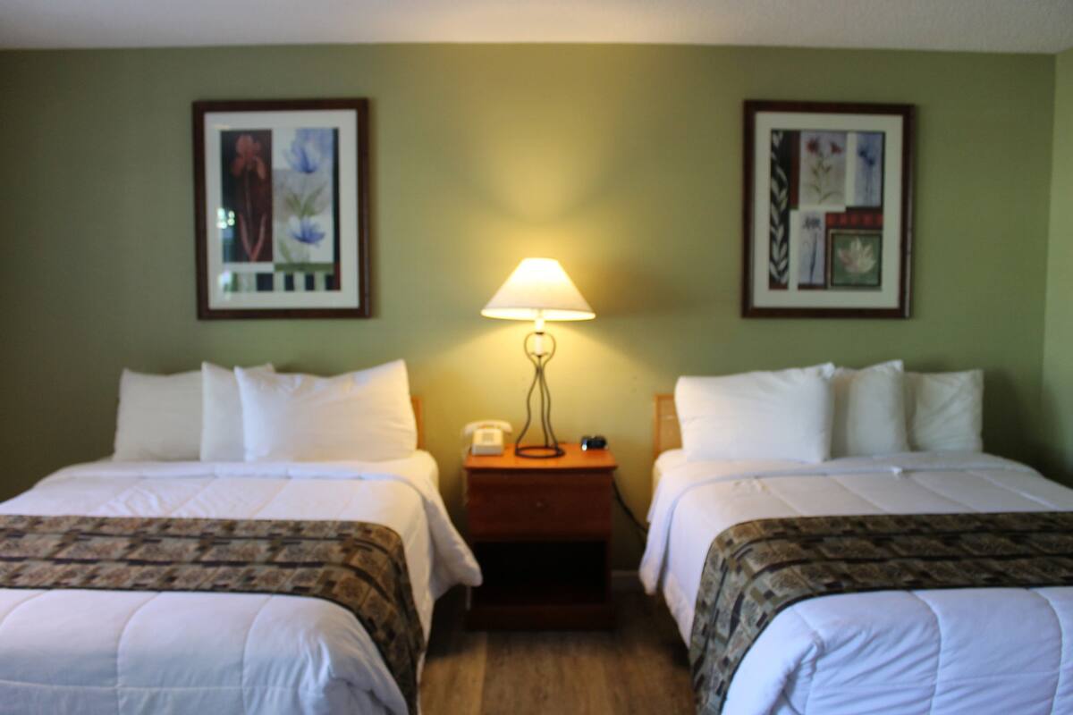 Quarto de hotel com duas camas, uma mesinha de cabeceira com abajur e quadros nas paredes.