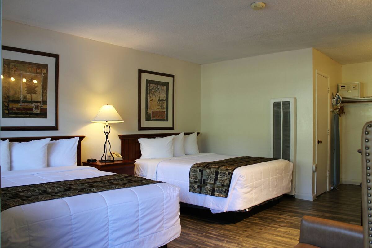 Quarto de hotel com duas camas, uma mesa de cabeceira com abajur, um quadro emoldurado na parede e uma porta com porta-toalhas.
