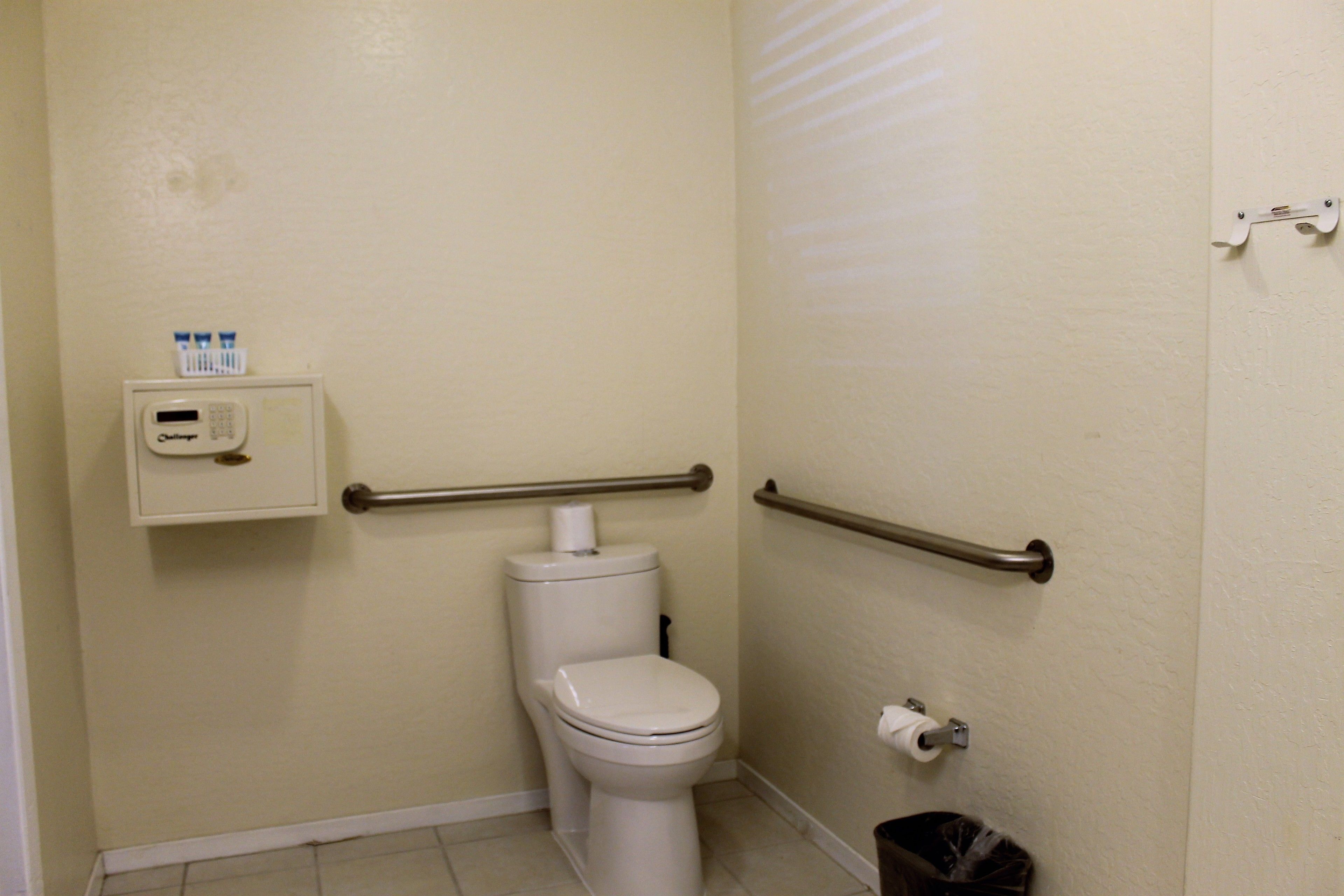 A small, clean bathroom with a toilet, handrails, a trash can, and a soap dispenser on the wall.
