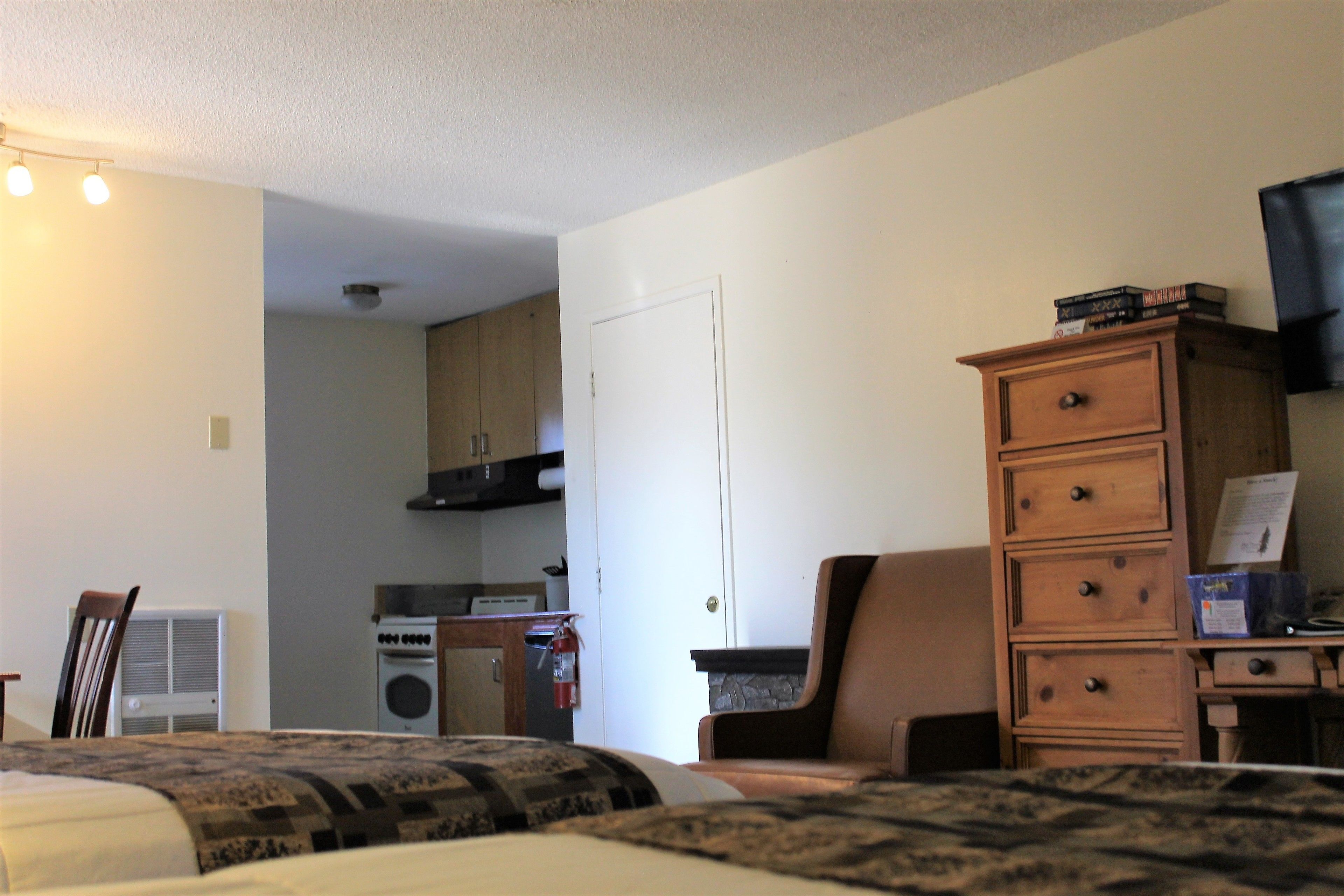 A hotel room with a bed, a dresser, a chair, a television, and a kitchenette in the background.