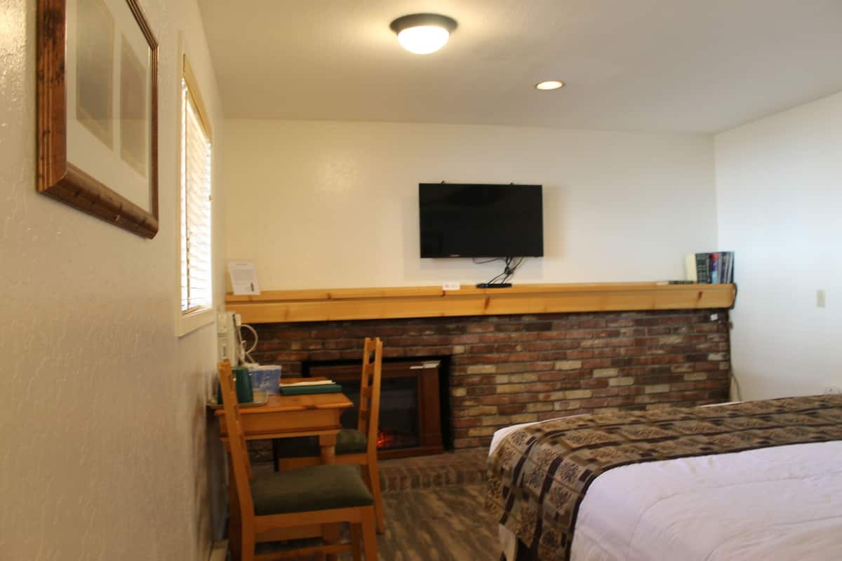 Quarto de hotel com lareira de tijolos, televisão presa na parede, uma cadeira de madeira e cama com colcha estampada.