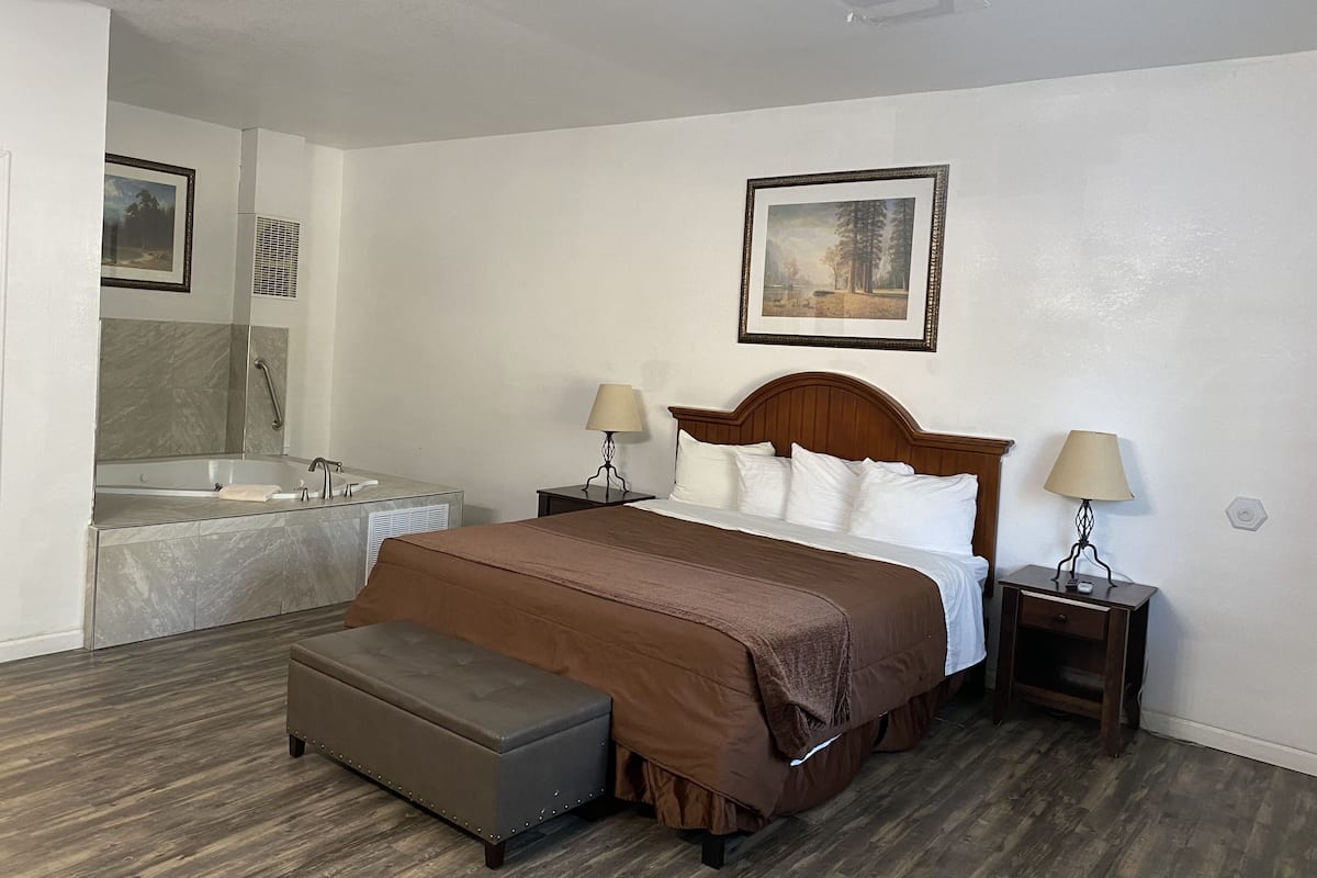 Quarto de hotel com uma cama grande, um banco, banheiro com banheira e duas mesas de cabeceira com luminárias.