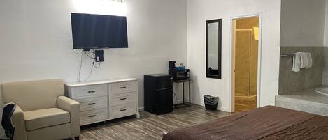 Quarto de hotel com cama, escrivaninha com computador, cadeira, penteadeira, televisão, espelho e banheiro com chuveiro e banheira.