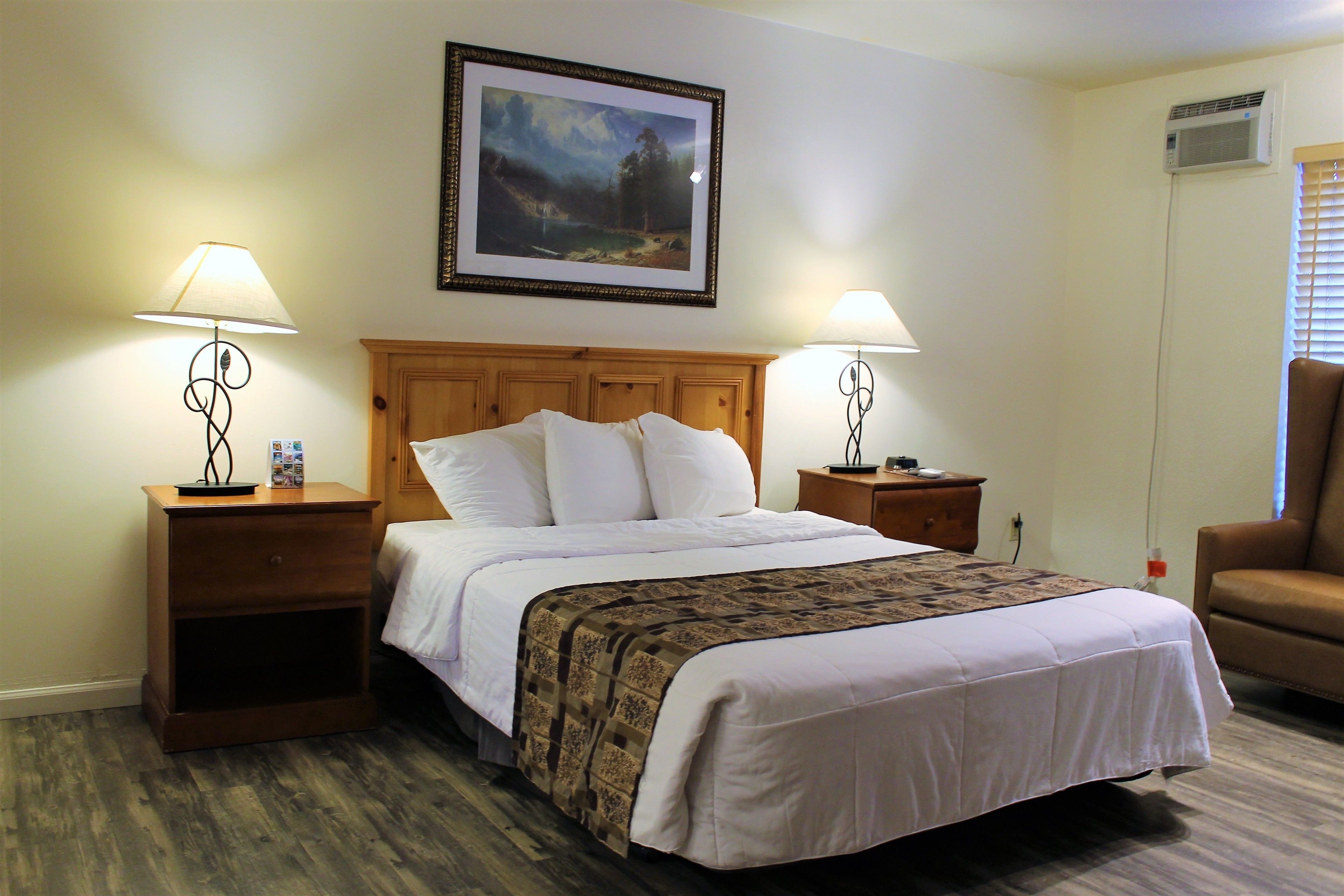 A hotel room with a large bed, two bedside tables with lamps, a wooden headboard, a framed picture on the wall, and a window with blinds.