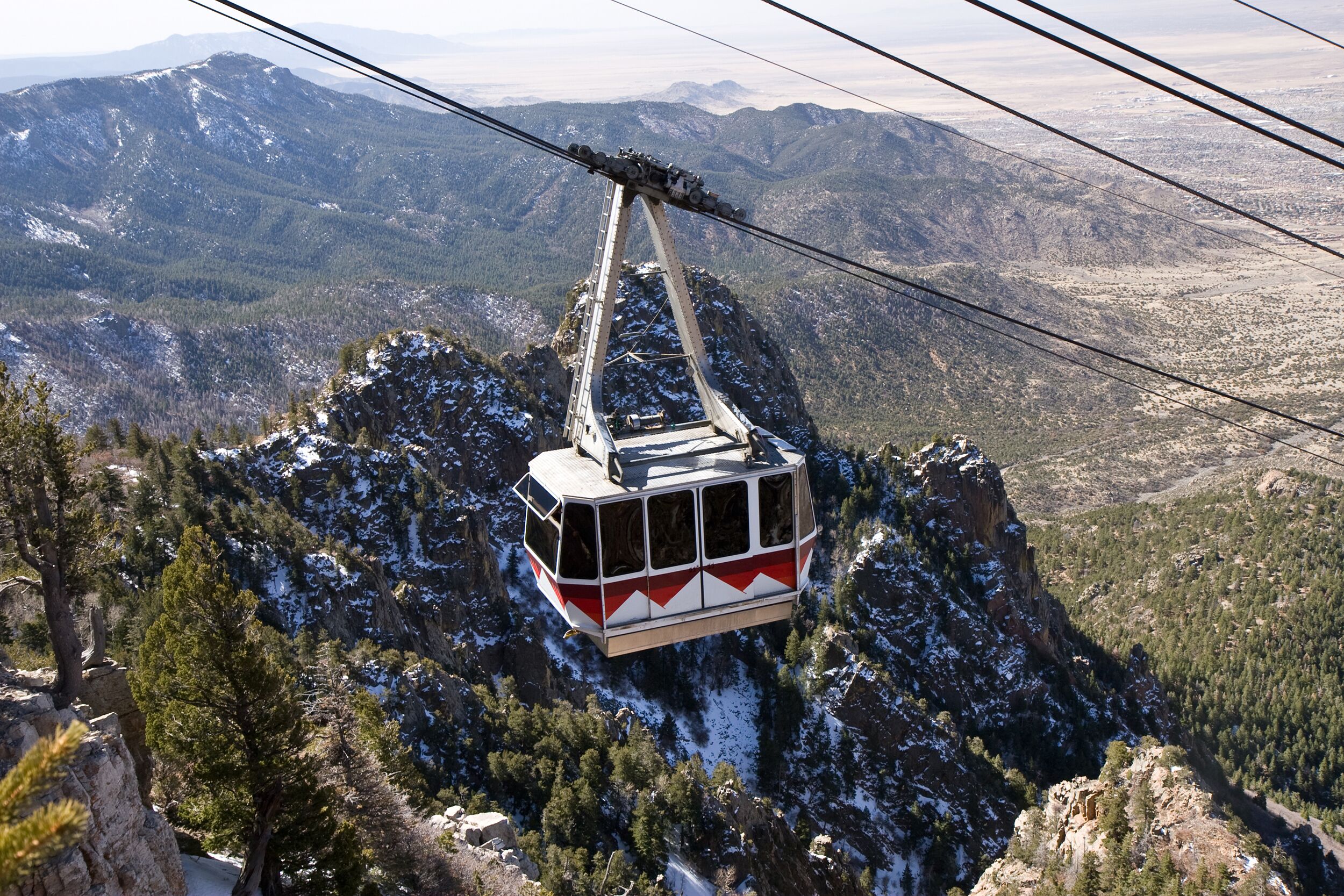 A cable car suspended over a mountainous landscape with snow-covered peaks and dense forests.