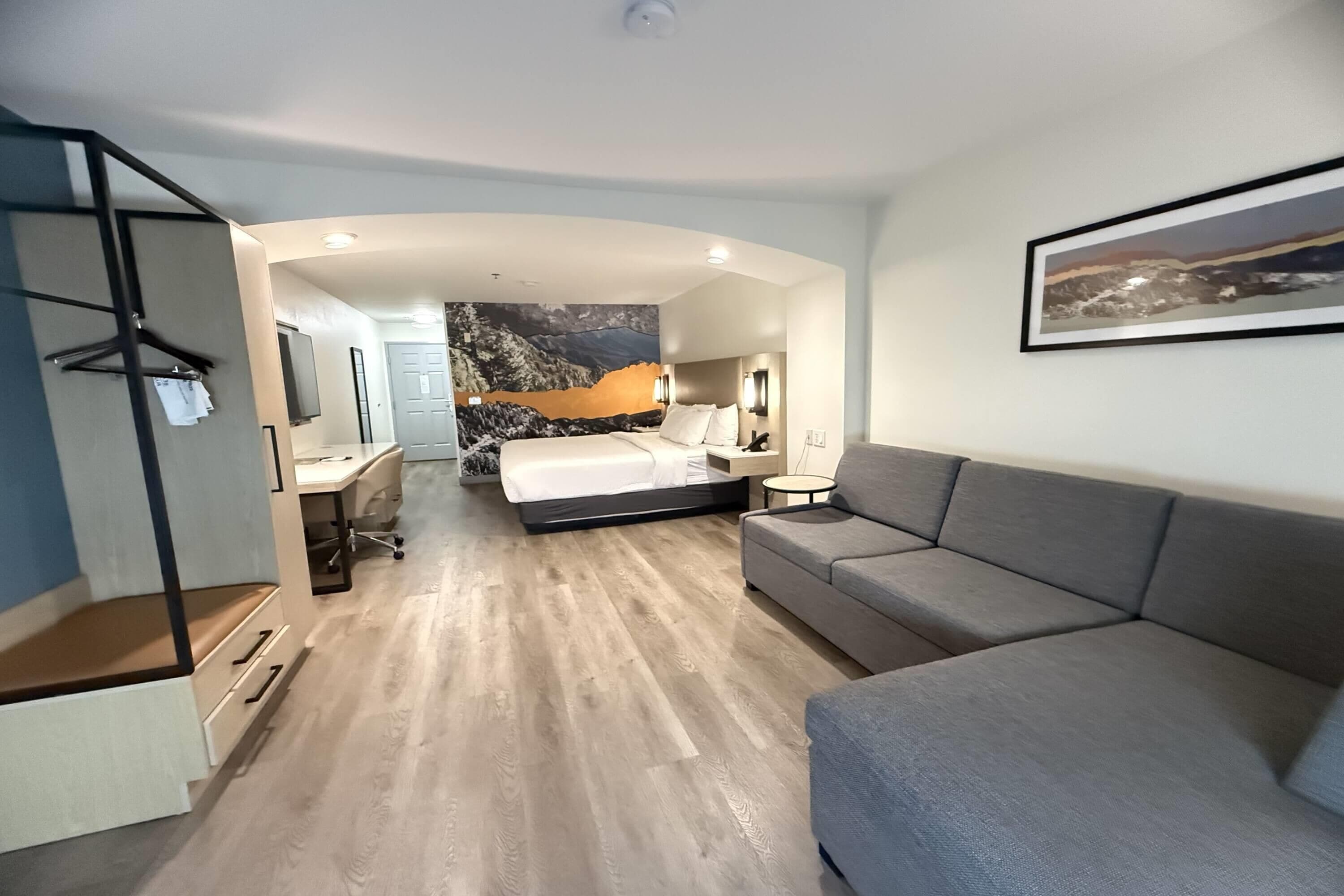 A modern hotel room with a bed, a grey sofa, a desk, and a chair.