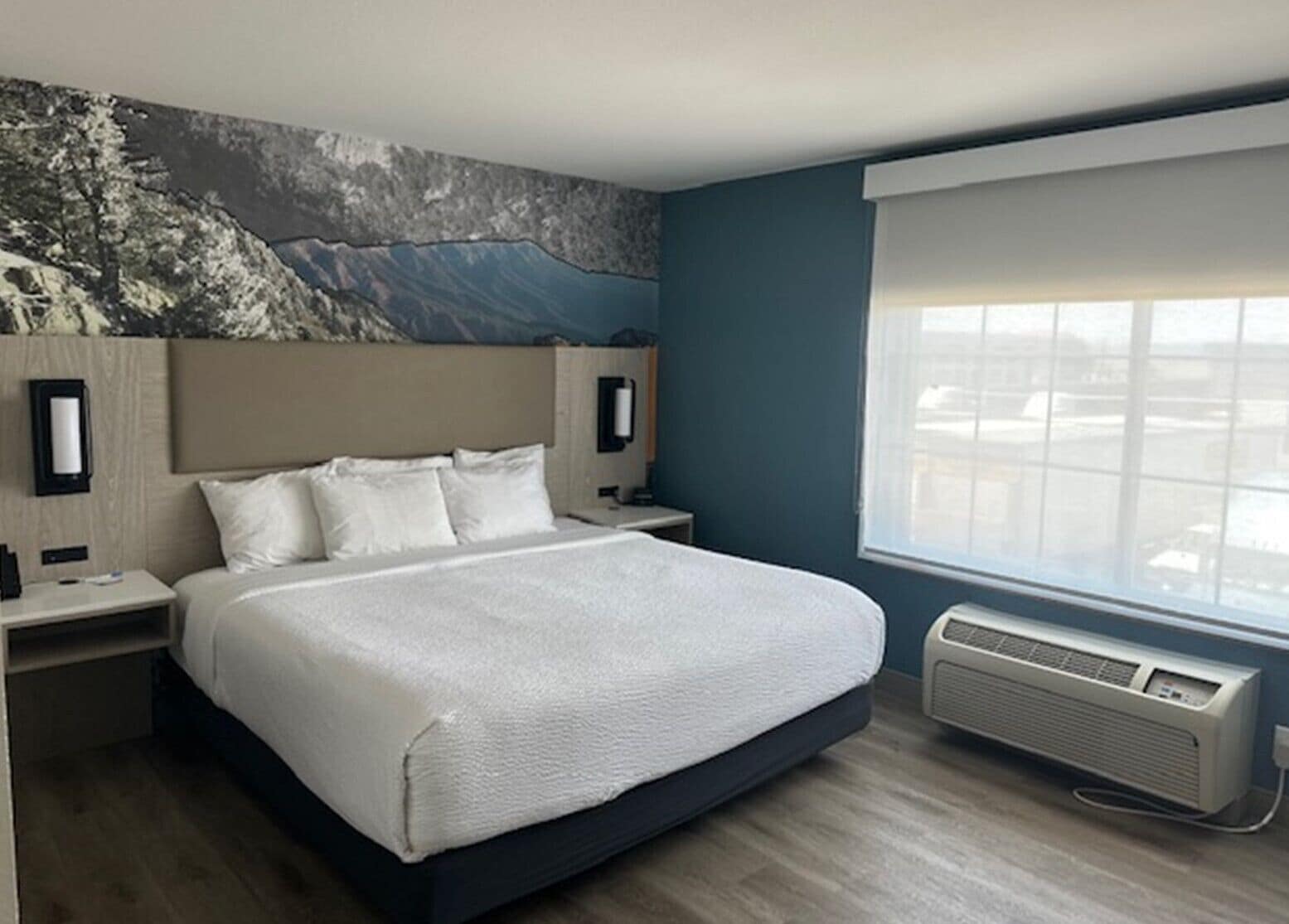 A modern hotel room with a large bed, a mounted artwork on the wall, a bedside table, and a window with a view.