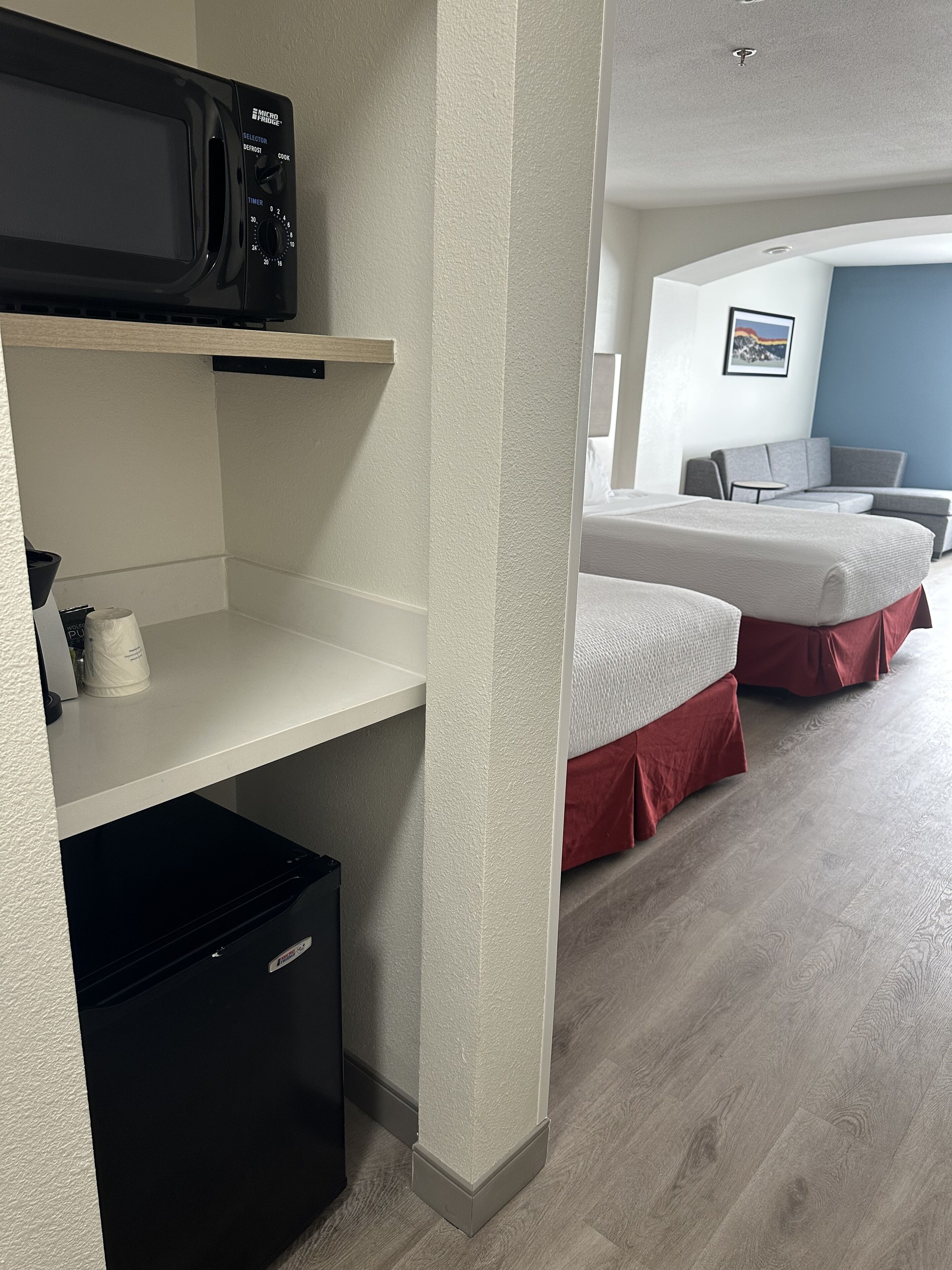 A hotel room with a kitchenette, a bed with red bedding, a desk, and a view of the cityscape.