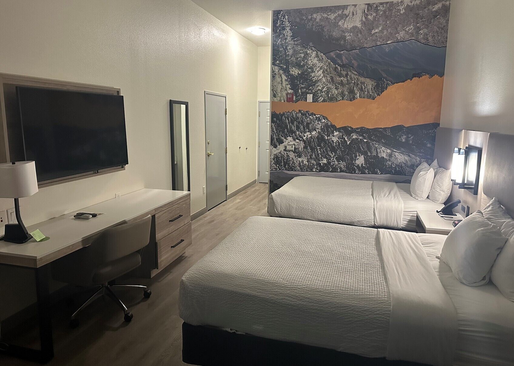 A hotel room with a desk, two beds, a large mountain painting, and a TV.
