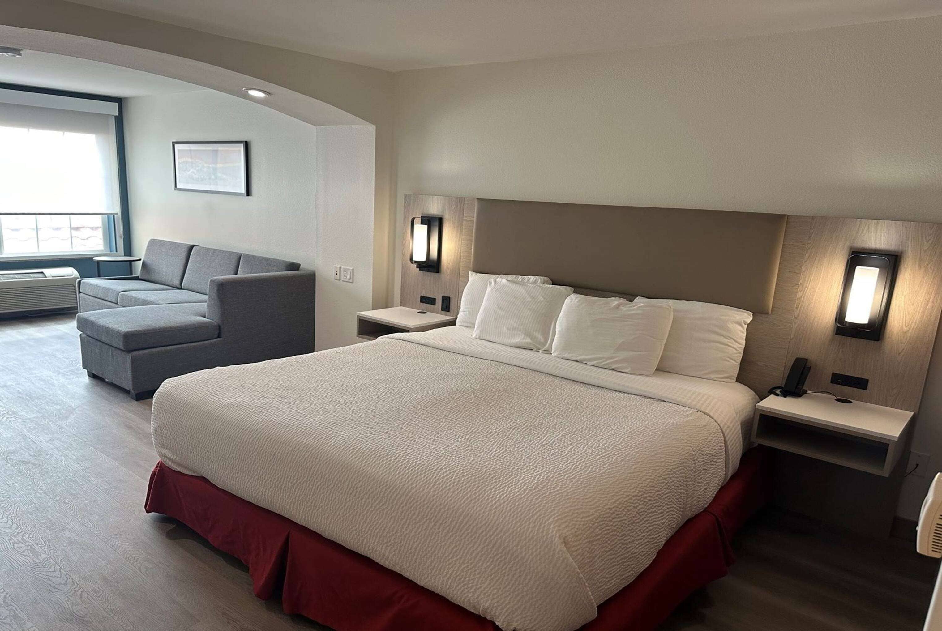 A hotel room with a large bed, a gray armchair, a small table, and a nightstand with a lamp.