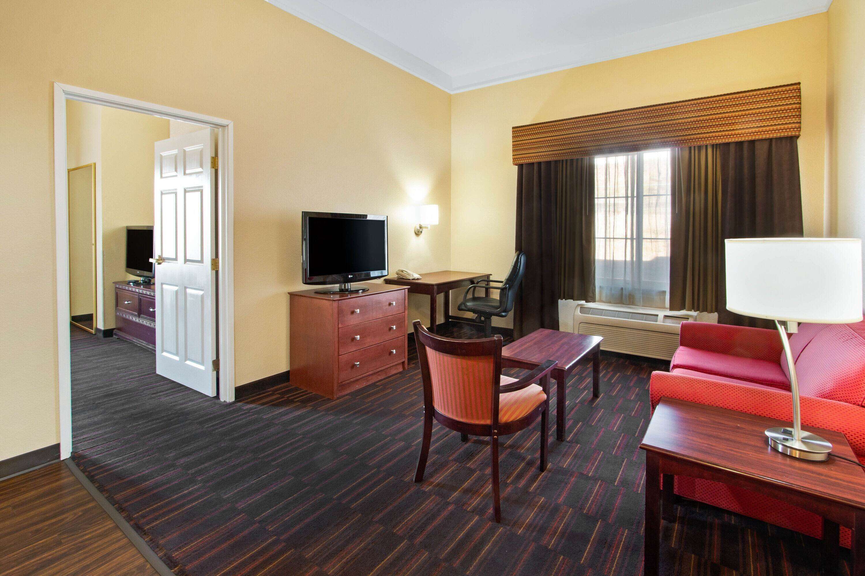 A hotel room with a TV, desk, chair, sofa, and a lamp.