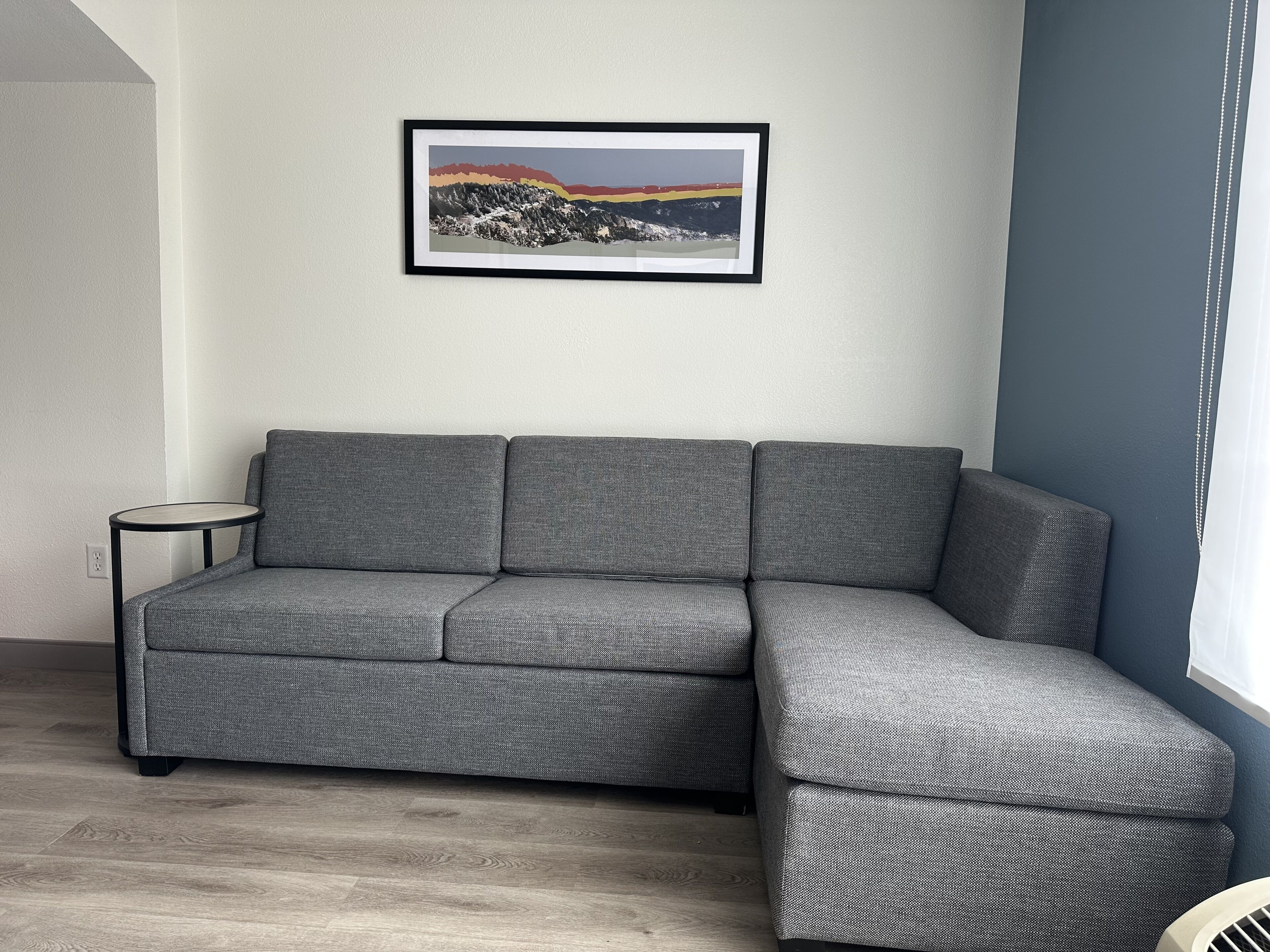 En grå modulsofa med et lille sofabord, et billedramme med et landskabsmotiv på væggen, og et vindue med gardiner.