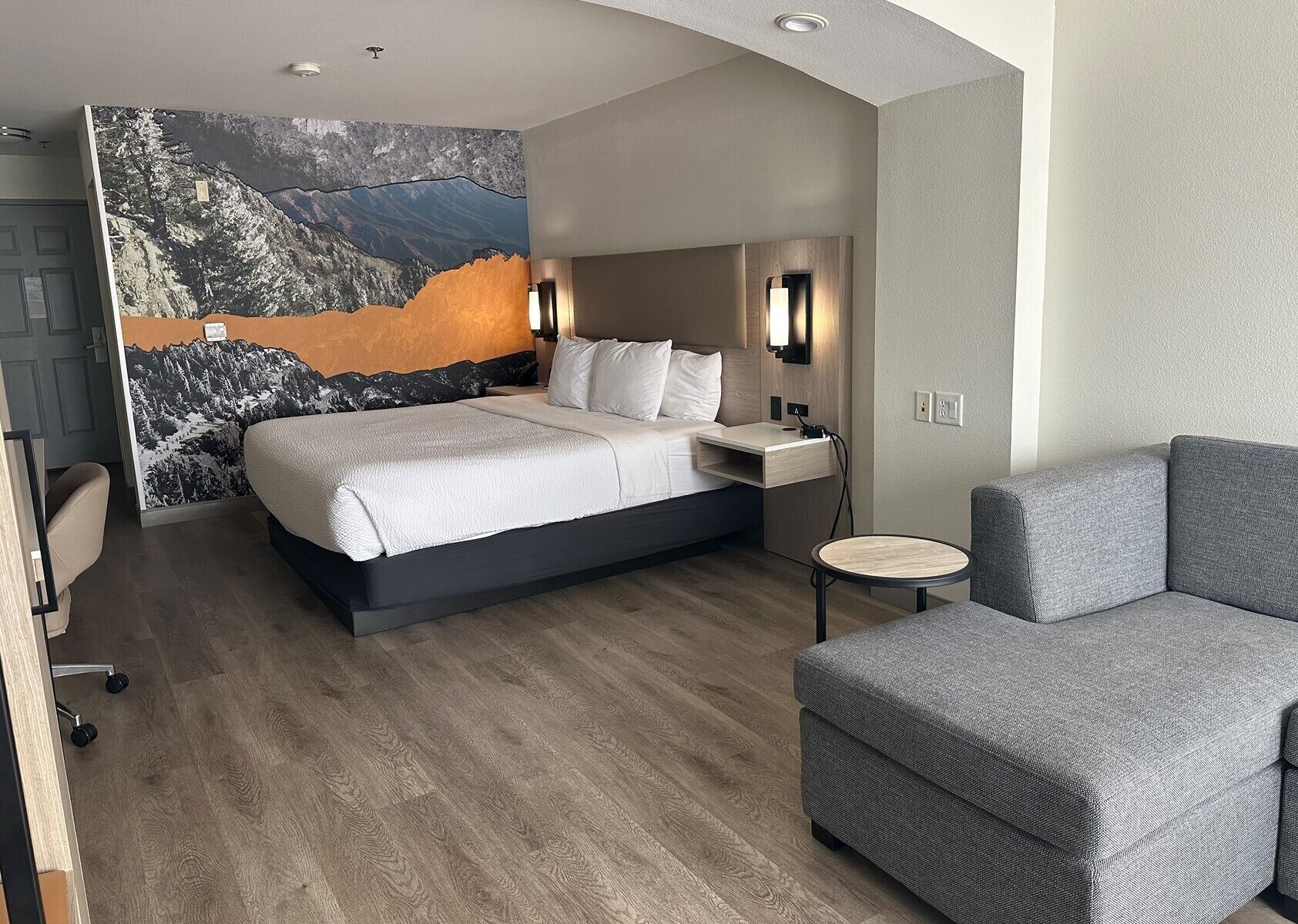 Una habitación de hotel moderna con una cama grande, un sofá gris, una mesita pequeña y un mural en la pared.