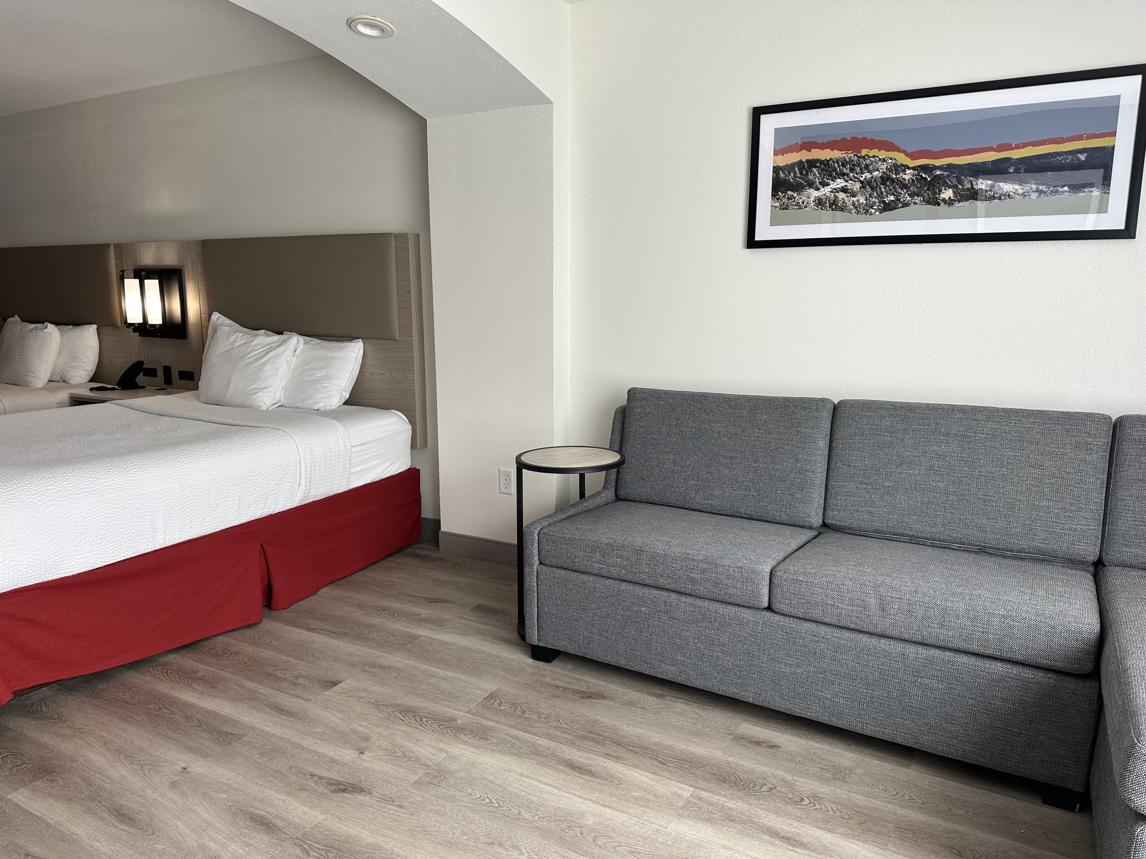 A hotel room with a bed, a grey sofa, a small round table, and a framed picture on the wall.