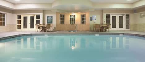 A swimming pool with a tiled deck, surrounded by doors and windows, and a few chairs.
