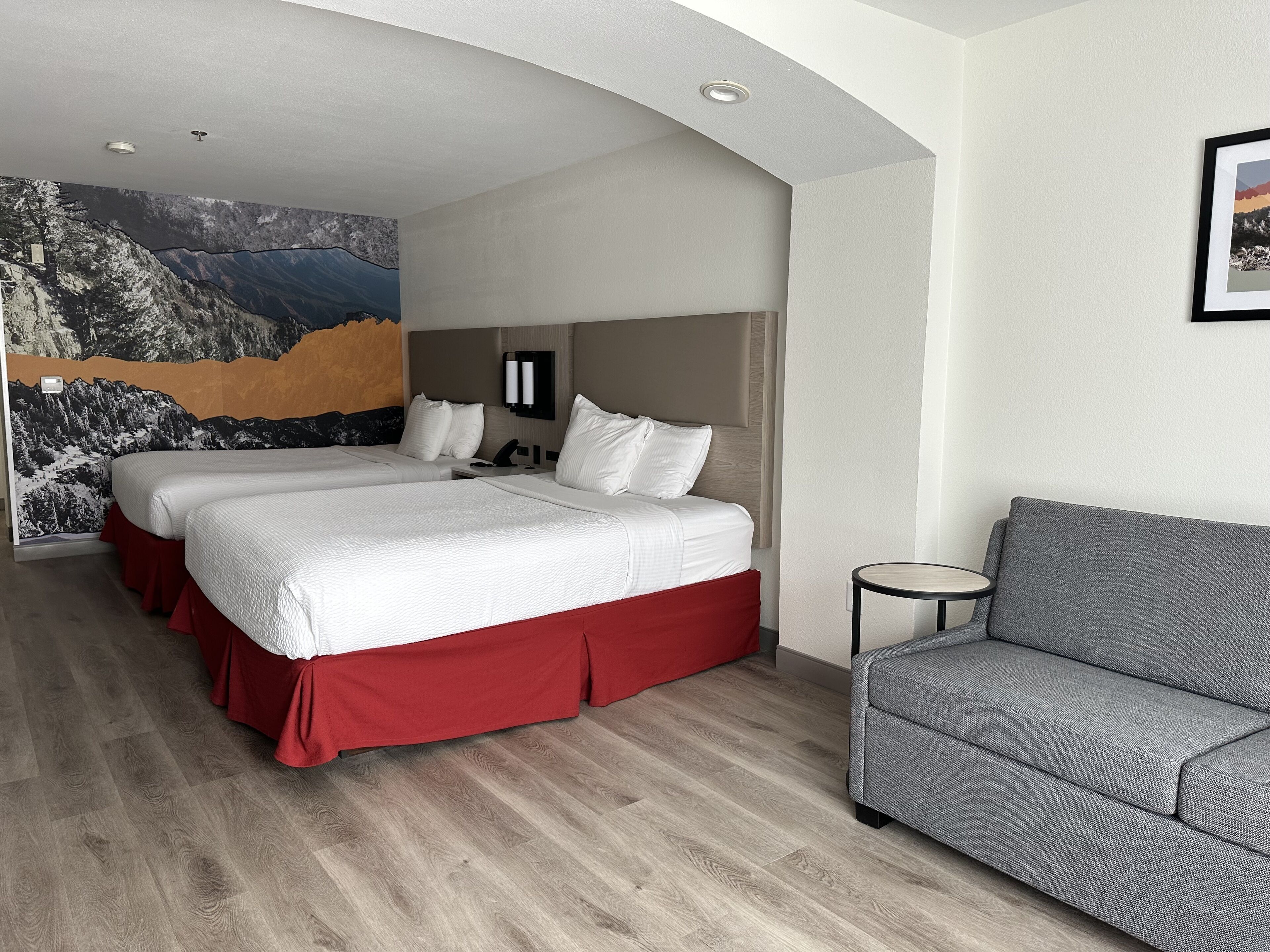 A hotel room with two beds, a gray sofa, a small table, and a mural on the wall.