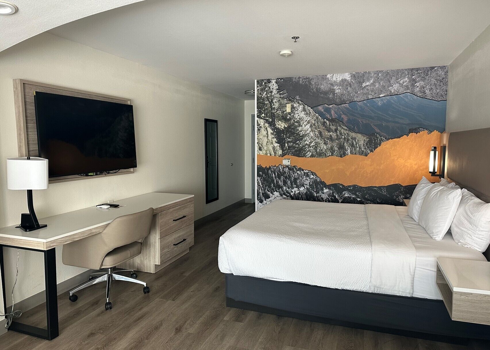 A hotel room with a bed, a desk, a chair, a TV, and a large mountain landscape painting on the wall.