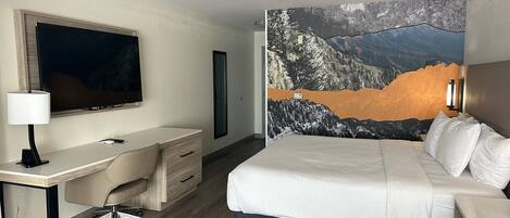 A hotel room with a bed, a desk, a chair, a TV, and a large mountain landscape painting on the wall.