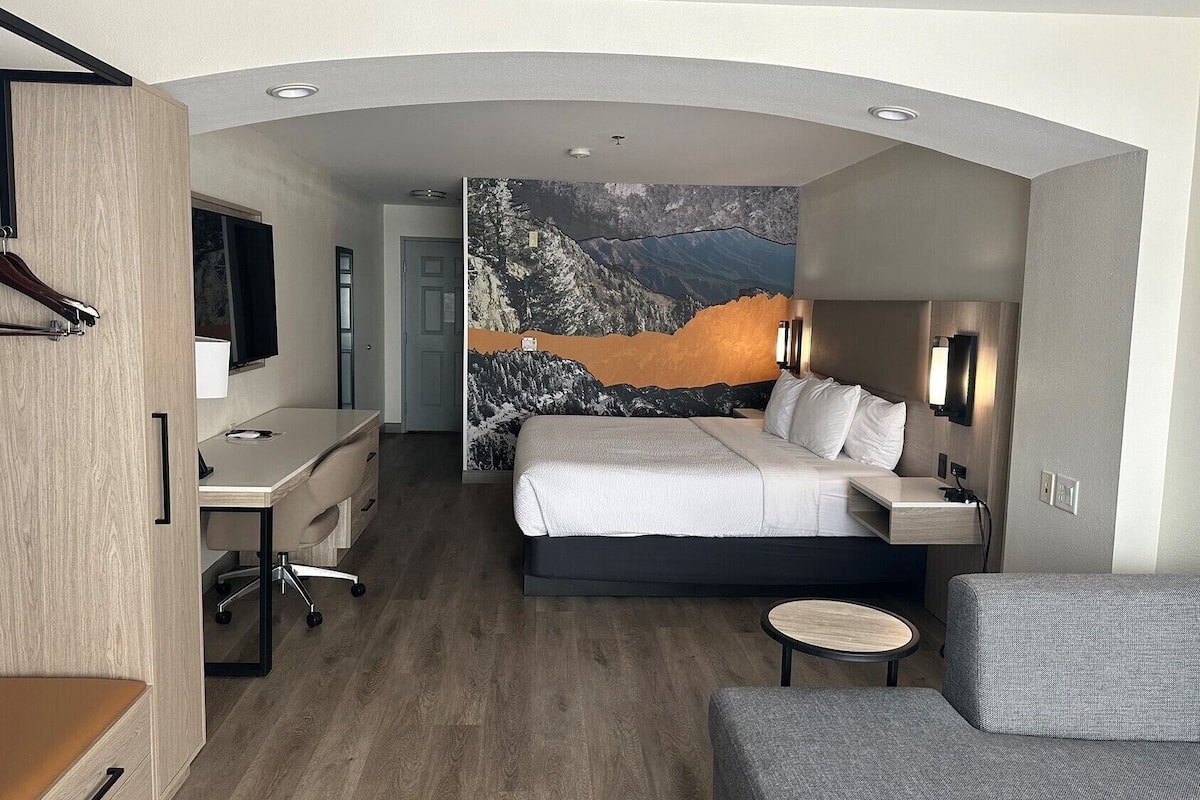 A modern hotel room with a large bed, a desk, a chair, and a small table.