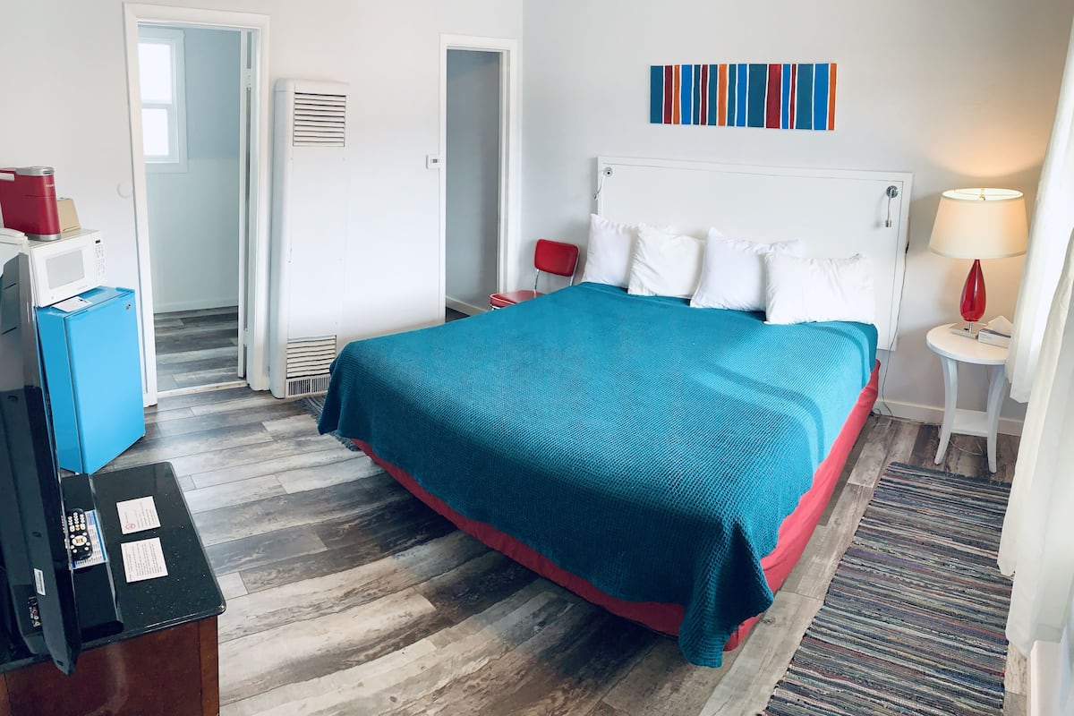 A hotel room with a bed, a desk, a chair, a microwave, a refrigerator, and a red chair.