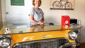 Un coche amarillo antiguo exhibido en una sala de exposición, con una mujer de pie detrás.