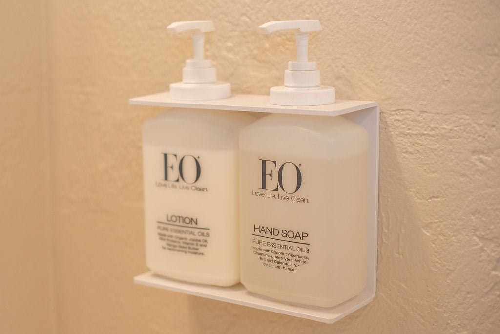 To flaske EO lotion og håndsæbe står på en hvid reol foran en væg med struktur.